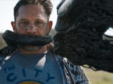 In dieser Szene aus Venom 3 ist Tom Hardy als Eddie Brock zu sehen, wie ihn der schwarze, flüssige Symbiont Venom teilweise umhüllt und seinen Mund bedeckt. Er trägt ein graues T-Shirt mit der Aufschrift „The City“ und blickt ernst nach vorne. Die Umgebung im Hintergrund ist verschwommen, was den Fokus auf die bedrohliche Verbindung zwischen Brock und Venom legt.