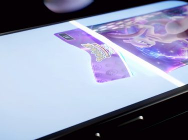 Auf dem Bild zu dem Artikel über die Pokémon TCG Pocket Tipps ist ein Smartphone-Display zu sehen, auf dem eine holografische Pokémon-Karte erscheint. Die Karte schimmert in violettem Licht, während das Logo von Pokémon Trading Card Game hervorgehoben ist. Das Smartphone liegt leicht angewinkelt auf einer dunklen Oberfläche, wodurch der futuristische Effekt der Darstellung betont wird.