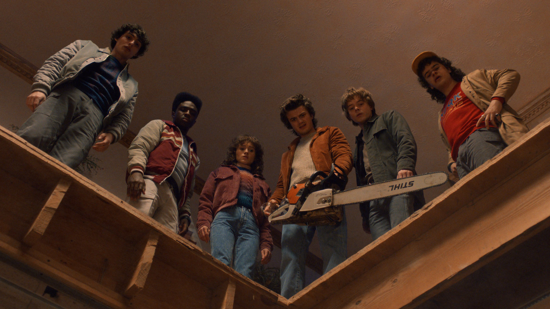 Auf diesem Bild aus Stranger Things Staffel 5 stehen sechs Charaktere in einer dramatischen Szene am Rand einer Holzöffnung und blicken angespannt nach unten. Von links nach rechts sind zu sehen: Finn Wolfhard als Mike, Caleb McLaughlin als Lucas, Natalia Dyer als Nancy, Joe Keery als Steve mit einer Kettensäge, Charlie Heaton als Jonathan und Gaten Matarazzo als Dustin. Die Gruppe wirkt konzentriert und entschlossen.