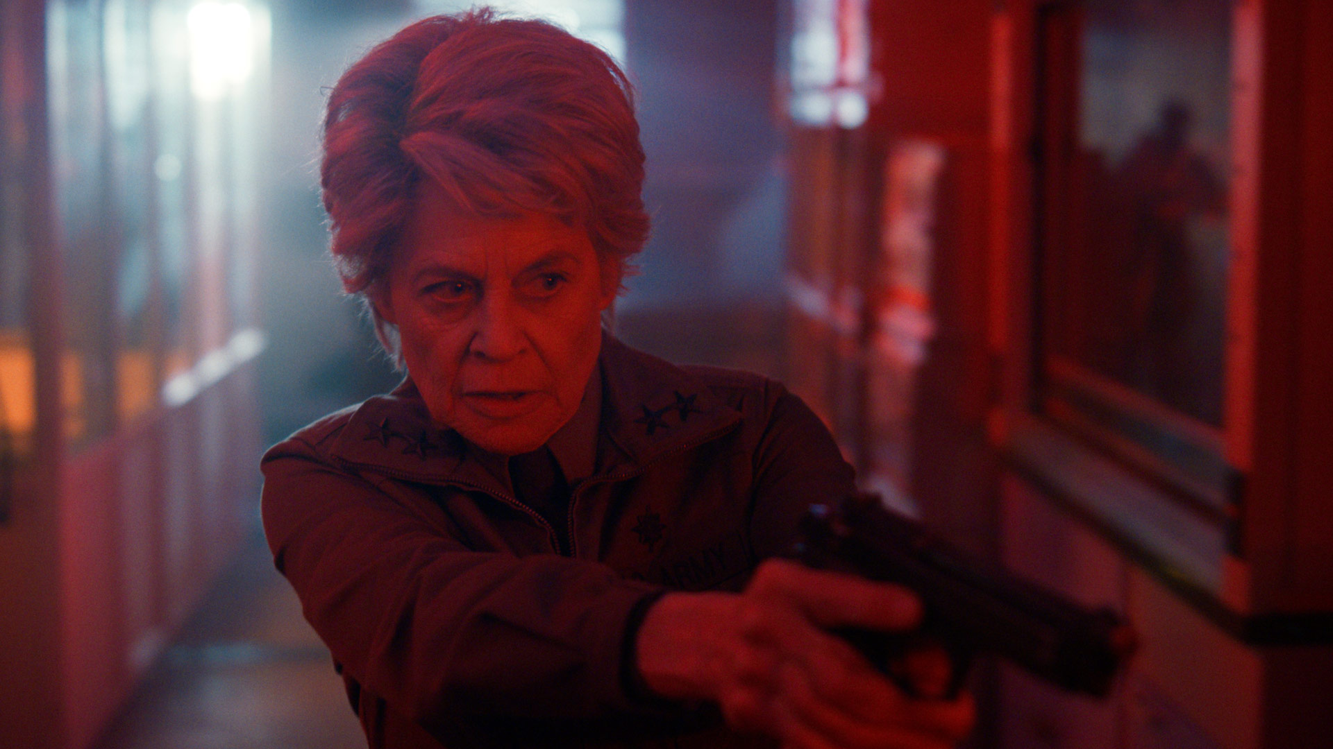 Auf dem Bild aus Stranger Things Staffel 5 ist Linda Hamilton als Dr. Kay zu sehen. Sie steht in einem rot beleuchteten Korridor, hält eine Pistole mit beiden Händen und blickt fokussiert und angespannt nach vorne. Ihre militärische Jacke und der entschlossene Gesichtsausdruck unterstreichen ihre kampferprobte und autoritäre Rolle.