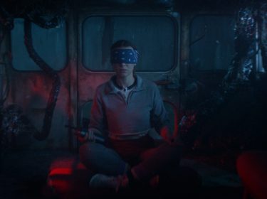 Auf diesem Bild aus Stranger Things Staffel 5 sitzt Millie Bobby Brown als Elf mit verschränkten Beinen in einem verlassenen Bus, der von dunklen Tentakeln überwuchert ist. Elf trägt eine dunkelblaue Augenbinde und hält ein Walkie-Talkie in der rechten Hand.