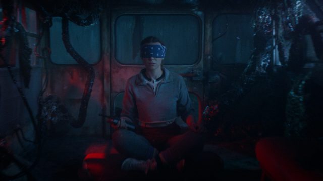 Auf diesem Bild aus Stranger Things Staffel 5 sitzt Millie Bobby Brown als Elf mit verschränkten Beinen in einem verlassenen Bus, der von dunklen Tentakeln überwuchert ist. Elf trägt eine dunkelblaue Augenbinde und hält ein Walkie-Talkie in der rechten Hand.