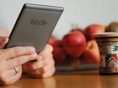 Hände halten einen Kindle-eReader. Auf dem Tisch steht eine bunte Tasse, im Hintergrund eine Obstschale mit Äpfeln gefüllt.