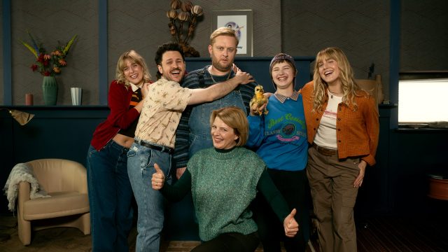 Auf dem Bild zur Serie Kacken an der Havel posieren die Schauspieler Anton "Fatoni" Schneider, Dimitrij Schaad, Sky Arndt, Jördis Triebel, Jana Münster und Sophia Münster gut gelaunt vor einer Wohnzimmerkulisse. Die Stimmung ist ausgelassen, viele lachen in die Kamera, während Dimitrij Schaad den ernsten Anton Schneider umarmt. Sky Arndt hält dabei ein kleines Stofftier in der Hand.