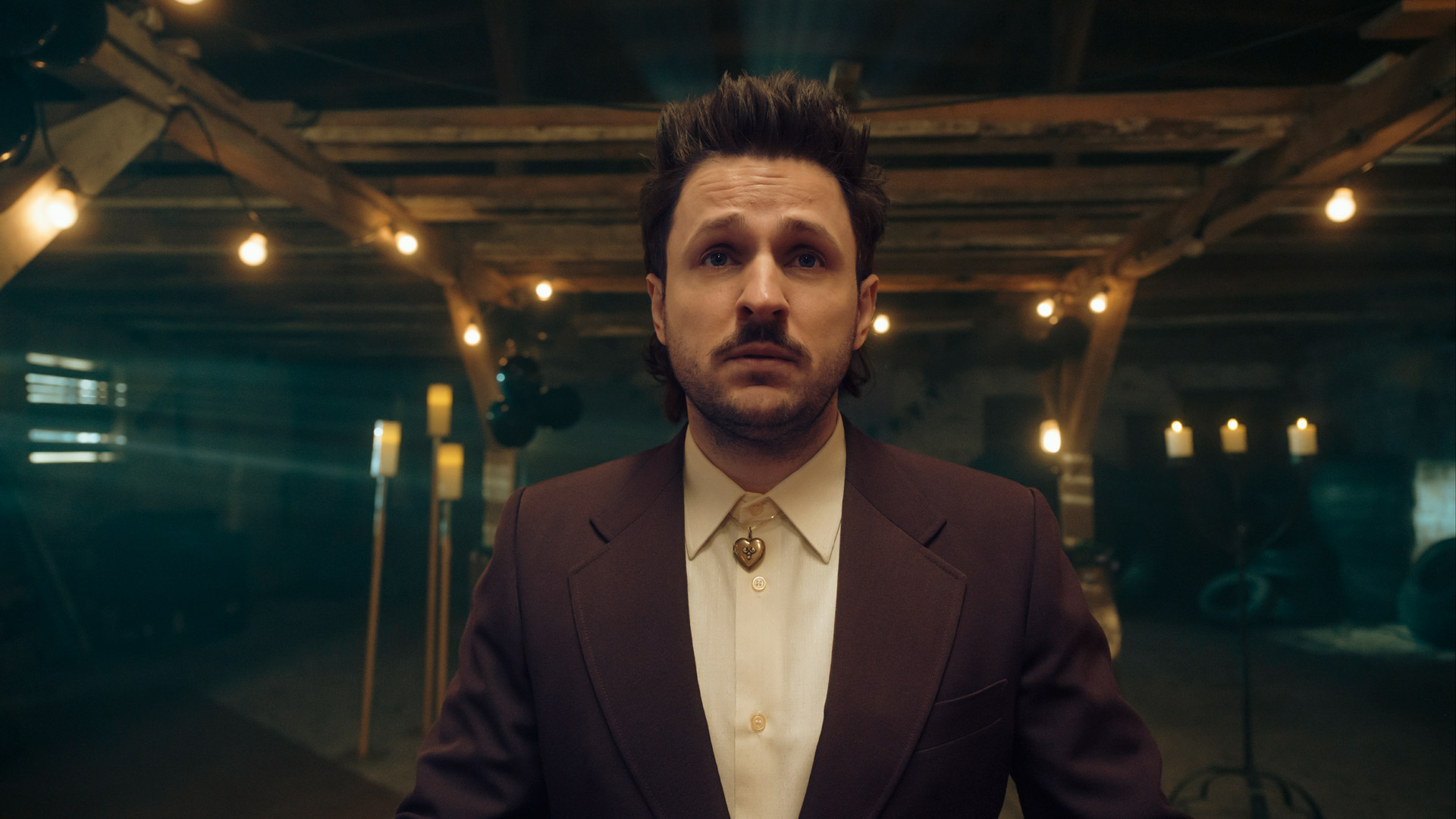 In dieser Szene aus Kacken an der Havel steht Dimitrij Schaad in einem dunklen, stimmungsvoll beleuchteten Raum mit Kerzen und Lichterketten. Er trägt einen weinroten Anzug und eine cremefarbene Hemdbluse mit einem auffälligen Herzanhänger. Sein ernster Blick verleiht der Szene eine emotionale, fast feierlich-melancholische Stimmung.