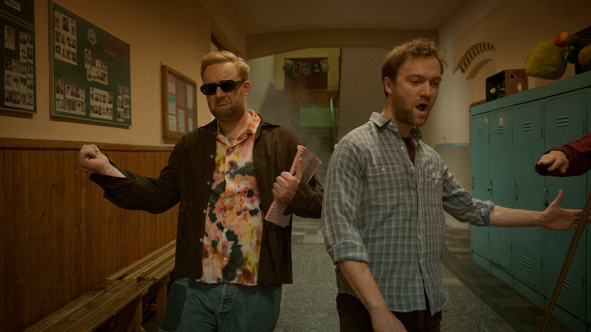 In einer Szene aus Kacken an der Havel gehen Anton „Fatoni“ Schneider und Vincent Redetzki durch einen Schulflur. Anton trägt Sonnenbrille und ein buntes Hemd, während er ein Notizbuch hält. Vincent schaut überrascht zur Seite. Die beiden wirken entschlossen und leicht überdreht, was den schrägen Humor von Kacken an der Havel unterstreicht.