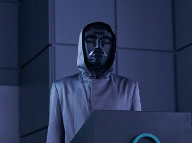 Das Bild im Artikel zum Squid Game US-Remake zeigt eine Szene aus der zweiten Staffel der Originalserie, in der der Frontman, eine mysteriöse Figur in einem kantigen, schwarzen, geometrischen Maske und einem grauen Kapuzenmantel, hinter einem Pult steht, umgeben von einer düsteren, blau beleuchteten Umgebung.