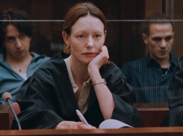 Auf dem Bild in dem Artikel über die Zeit Verbrechen-Serie, ist eine Frau mit roten Haaren zu sehen, die wie eine Anwältin gekleidet ist. Im Hintergrund sind weitere Menschen zu sehen. Der Raum in dem sie sitzen scheint ein Gerichtssaal zu sein.