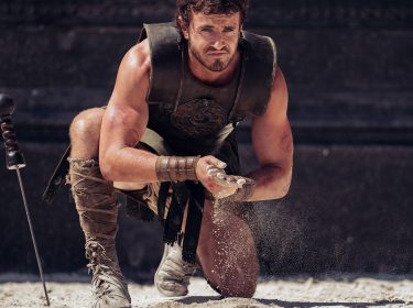 Dieses Bild aus Gladiator 2 zeigt Lucius, gespielt von Paul Mescal, in einer intensiven Kampfszene. Gekleidet in Gladiatorenrüstung, kniet er mit konzentriertem Blick und streut symbolisch Sand auf den Boden, was seine Entschlossenheit und die Vorbereitung auf einen bevorstehenden Kampf ausdrückt. Neben ihm steckt ein Schwert im Boden, das auf seine Rolle als Kämpfer hinweist. Die Szene ist dramatisch beleuchtet und betont die körperliche und mentale Stärke des Charakters in der Arena.