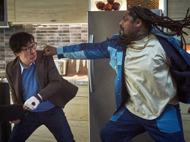 In einer Szene aus Love Hurts - Liebe tut weh ist links Ke Huy Quan zu sehen, der sichtlich verletzt wirkt und einen Laptop hält. Rechts steht Marshawn Lynch, der in dynamischer Bewegung mit ausgestrecktem Arm zum Schlag ansetzt. Die Szene spielt in einer modernen Küche, deren Details wie ein Kühlschrank und eine geflieste Wand im Hintergrund zu erkennen sind.