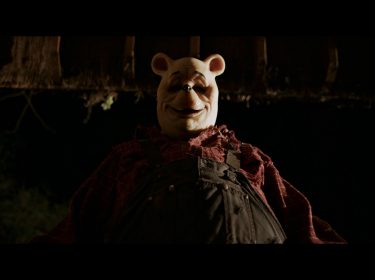 Dieses Bild aus dem Film Winnie the Pooh: Blood and Honey zeigt eine düstere, unheimliche Version von Winnie Puuh, die das klassische Kinderbuch-Image ins Horror-Genre überführt. Die Figur trägt eine beunruhigende Maske mit markanten, kindlichen Zügen, kombiniert mit abgenutzter Latzhose und kariertem Hemd. Die Szene ist in dunklen, bedrohlichen Farben gehalten, was die gruselige Atmosphäre unterstreicht. Der Film ist der erste in einer Reihe von Kinderbuch-Horrorfilmen im Poohniverse.