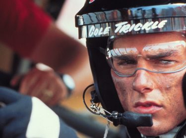 Auf dem Bild aus Tage des Donners 2 ist Cole Trickle, gespielt von Tom Cruise, in Nahaufnahme zu sehen. Er trägt einen Rennhelm mit seinem Namen darauf und eine Schutzbrille, sein Gesicht ist verschmutzt von Rennstaub. Die Szene vermittelt Spannung und Konzentration, während er sich auf das Rennen vorbereitet.