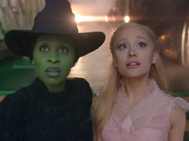 Auf dem Bild aus dem Wicked-Film sind Cynthia Erivo als Elphaba und Ariana Grande als Glinda zu sehen. Elphaba hat grüne Haut, trägt ein schwarzes Kleid und einen spitzen Hut, während Glinda in einem zarten rosa Kleid mit verträumtem Blick dargestellt ist. Beide schauen fasziniert in die Ferne, umgeben von einer mystischen und leuchtenden Atmosphäre.