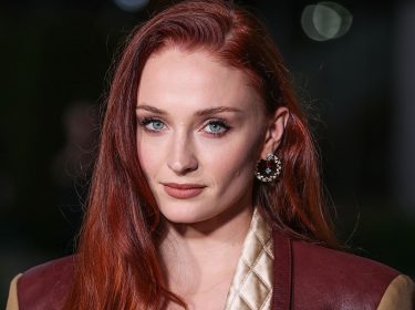 Auf dem Pressebild zur Tomb Raider-Serie auf Amazon Prime Video ist Sophie Turner zu sehen, die als mögliche Hauptdarstellerin gehandelt wird. Sie trägt ihr rotes Haar offen, dezentes Make-up und auffällige Ohrringe. Ihr Look wird durch eine gesteppte, beige Jacke mit braunen Lederakzenten ergänzt. Der Hintergrund ist dunkel mit weichen Lichtreflexen.