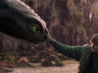 Auf dem Bild zur neuen Verfilmung von Drachenzähmen leicht gemacht für 2025 ist der schwarze Drache Ohnezahn mit grünen Augen links zu sehen, wie er sanft mit Hicks interagiert. Hicks, rechts im Bild, trägt grüne Kleidung mit einer Fellweste und berührt die Nase von Ohnezahn. Die Szene spielt in einer naturbelassenen, waldigen Umgebung mit gedämpften Farben, die eine ruhige und emotionale Atmosphäre schaffen.