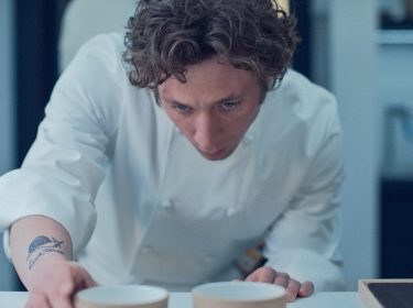 Auf dem Bild zur Serie The Bear Staffel 4 ist Carmen „Carmy“ Berzatto, gespielt von Jeremy Allen White, zu sehen. Er trägt eine weiße Kochjacke und ist konzentriert, während er zwei Schalen auf einem Tisch präzise arrangiert. Sein Blick ist fokussiert, und ein Schnecken-Tattoo ist auf seinem Unterarm sichtbar. Die Szene vermittelt eine intensive Arbeitsatmosphäre in einer professionellen Küche.