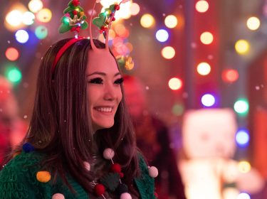 Im Weihnachtsspecial von Guardians of the Galaxy ist Mantis zu sehen. Sie trägt ein festliches Stirnband mit kleinen Weihnachtsbäumen und bunte Dekoration an ihrem grünen Pullover. Mit einem fröhlichen Lächeln steht sie inmitten einer weihnachtlichen Kulisse mit bunten Lichtern und fallendem Schnee, was die festliche Stimmung unterstreicht.