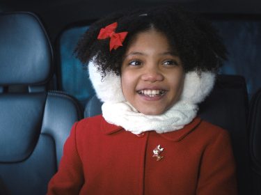 Auf dem Bild im GigaTV-Highlights Dezember 2024-Artikel siehst Du Leah Brady als Trudy in Violent Night. Sie sitzt lächelnd in einem Auto, trägt eine rote Jacke mit einer Rentier-Brosche, einen weißen Schal und Ohrenschützer.