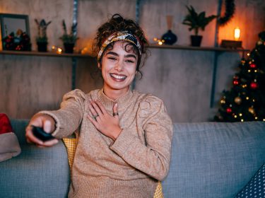 Eine lachende Frau sitzt gemütlich auf einem Sofa und schaut Weihnachtsepisoden von Serien
