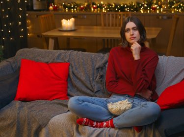 Eine junge Frau in einem roten Pullover sitzt auf einem grauen Sofa, umgeben von roten Kissen, und hält eine Schüssel Popcorn. Sie wirkt nachdenklich und leicht gelangweilt. Im Hintergrund ist ein festlich geschmückter Weihnachtsbaum mit Lichtern und roten Kugeln zu sehen, zusammen mit Kerzen und Lichterketten. Perfekt passend zum Thema 