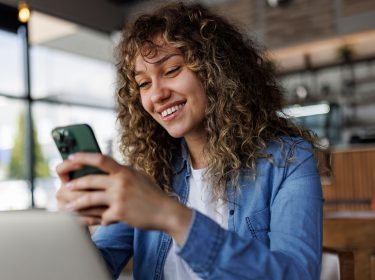 Eine junge Frau sitzt mit einem Handy in einem Café und lächelt.