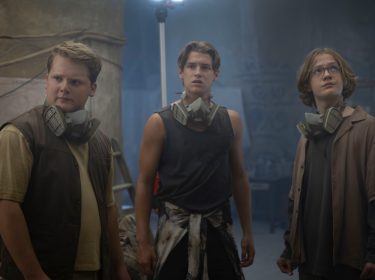 In „Die drei ??? – Toteninsel“ stehen Justus Jonas (Julius Weckauf), Peter Shaw (Nevio Wendt) und Bob Andrews (Levi Brandl) in einem staubigen, mysteriös beleuchteten Raum. Sie tragen alle Atemschutzmasken um den Hals und wirken alarmiert, während sie sich aufmerksam umsehen.