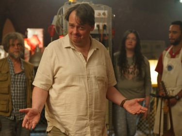 Auf dem Bild aus der Serie auf Amazon Prime Video ist Sascha Nathan in der Rolle des Gerry Star zu sehen. Gerry steht im Mittelpunkt einer Gruppe von vier Personen, die ihn aus dem Hintergrund beobachten. Er trägt ein beiges Hemd und blickt mit einem schelmischen Lächeln in die Kamera, seine Arme lässig ausgebreitet. Im Hintergrund sind Laborgeräte und eine industrielle Umgebung zu erkennen. Die Szene wirkt humorvoll und leicht chaotisch.