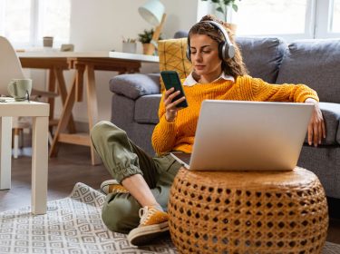 Frau mit Kopfhörern sitzt auf dem Boden vor einer Couch und hört Musik via Smartphone.