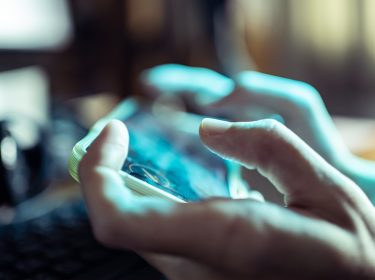 Das Bild in dem Artikel über die besten Koop-Games fürs Handy, zeigt eine Nahaufnahme von Händen, die ein Smartphone im Querformat halten. Der Fokus liegt auf den Fingern und dem Bildschirm, der unscharf, aber lebendig wirkt. Die Atmosphäre ist konzentriert, das Spielgeschehen spiegelt sich leicht auf dem Display wider.