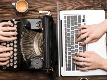 Das Bild zeigt einen Vergleich zwischen alt und neu: Links wird auf einer klassischen Schreibmaschine, einem typischen Retro-Gadget, getippt, während rechts ein moderner Laptop genutzt wird. Beide Geräte stehen nebeneinander auf einem Holztisch, was den Kontrast zwischen analogem und digitalem Arbeiten verdeutlicht.