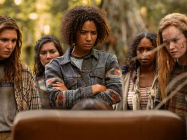 Auf dem Bild zu Yellowjackets Staffel 3 stehen fünf junge Frauen in einer Waldumgebung und blicken ernst auf etwas außerhalb des Bildes. Sophie Nélisse als Shauna, Jasmin Savoy Brown als Taissa, Liv Hewson als Van und andere Charaktere wirken angespannt und besorgt. Ihre Kleidung ist abgenutzt, ihre Gesichter zeigen Schmutz und Narben, was auf die harten Überlebensbedingungen hindeutet. Die Szene strahlt Spannung und Dramatik aus.