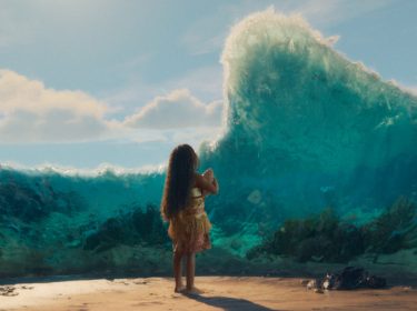 In dieser Szene aus der Vaiana-Realverfilmung steht ein kleines Mädchen mit langen, offenen Haaren barfuß am Strand und blickt auf eine gewaltige Wasserwand, die sich vor ihr auftürmt.