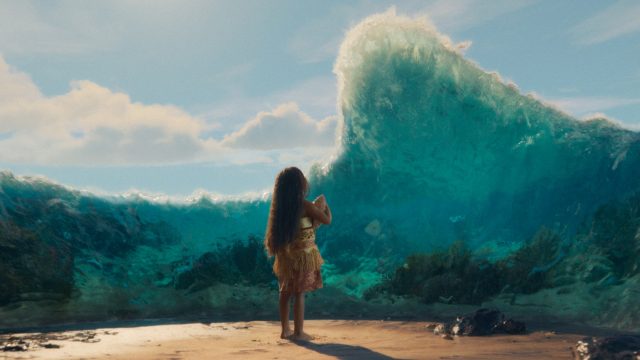 In dieser Szene aus der Vaiana-Realverfilmung steht ein kleines Mädchen mit langen, offenen Haaren barfuß am Strand und blickt auf eine gewaltige Wasserwand, die sich vor ihr auftürmt.