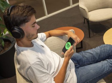 Junger Mann mit Kopfhörern sitzt auf Couch und hört am Smartphone Musik via Spotify