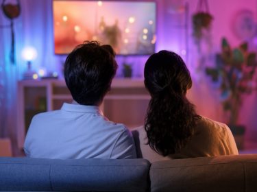 Auf dem Bild in dem Artikel zu den besten Silvester-Folgen in Serien, sitzen zwei Personen auf einem Sofa und schauen gemeinsam auf einen leuchtenden Fernseher, der eine warme Atmosphäre erzeugt. Der Raum ist mit bunten Lichtern dekoriert, passend zu einer festlichen Stimmung. Pflanzen und Regaldekorationen ergänzen die gemütliche Umgebung.