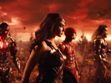 In dieser Szene aus dem DC Universum ist Gal Gadot als Wonder Woman im Vordergrund zu sehen, in ihrer ikonischen goldenen Rüstung. Links von ihr steht ein Cyborg-ähnlicher Charakter, während Jason Momoa als Aquaman ganz rechts mit seinem Dreizack abgebildet ist. Im Hintergrund erscheint ein Charakter in rotem Anzug. Der Himmel ist in dramatischen Rottönen gehalten und vermittelt eine bedrohliche Stimmung.