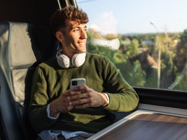 Ein Mann sitzt mit einem Smartphone und Kopfhörern im Zug und schaut aus dem Fenster
