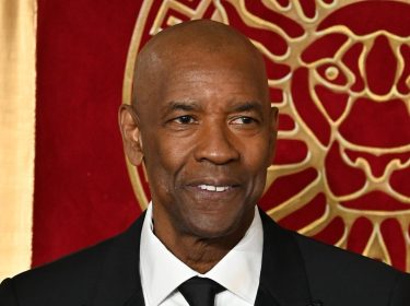 Auf dem Bild zu Denzel Washingtons besten Filmmomenten ist Denzel Washington in einem eleganten schwarzen Anzug mit weißem Hemd und schwarzer Krawatte zu sehen. Er lächelt dezent, während er vor einem roten Hintergrund mit goldenen Verzierungen, die ein Löwenmotiv zeigen, steht. Die Szene strahlt eine feierliche und stilvolle Atmosphäre aus.