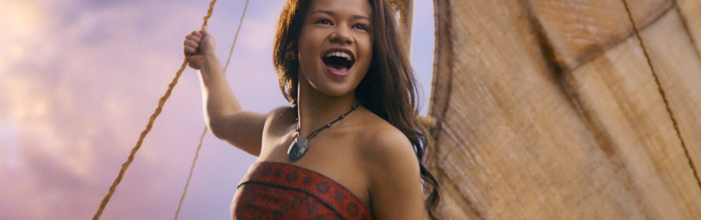 In der Szene aus den Disney Realverfilmungen 2026 sieht man Catherine Laga'aia als Vaiana auf einem traditionellen Segelboot bei Sonnenuntergang. Sie trägt ein rotes, gemustertes Oberteil und eine grüne Muschelkette. Mit offenem Mund und freudigem Blick hält sie sich an einem Seil fest, während das Segel im Hintergrund sichtbar ist. Die Stimmung wirkt abenteuerlich und optimistisch.