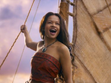 In der Szene aus den Disney Realverfilmungen 2026 sieht man Catherine Laga'aia als Vaiana auf einem traditionellen Segelboot bei Sonnenuntergang. Sie trägt ein rotes, gemustertes Oberteil und eine grüne Muschelkette. Mit offenem Mund und freudigem Blick hält sie sich an einem Seil fest, während das Segel im Hintergrund sichtbar ist. Die Stimmung wirkt abenteuerlich und optimistisch.