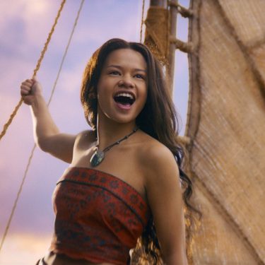In der Szene aus den Disney Realverfilmungen 2026 sieht man Catherine Laga'aia als Vaiana auf einem traditionellen Segelboot bei Sonnenuntergang. Sie trägt ein rotes, gemustertes Oberteil und eine grüne Muschelkette. Mit offenem Mund und freudigem Blick hält sie sich an einem Seil fest, während das Segel im Hintergrund sichtbar ist. Die Stimmung wirkt abenteuerlich und optimistisch.