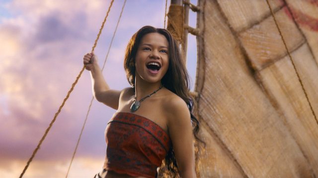 In der Szene aus den Disney Realverfilmungen 2026 sieht man Catherine Laga'aia als Vaiana auf einem traditionellen Segelboot bei Sonnenuntergang. Sie trägt ein rotes, gemustertes Oberteil und eine grüne Muschelkette. Mit offenem Mund und freudigem Blick hält sie sich an einem Seil fest, während das Segel im Hintergrund sichtbar ist. Die Stimmung wirkt abenteuerlich und optimistisch.