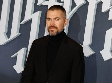 Auf dem Bild ist der Filmemacher Robert Eggers zu sehen, bekannt für seine atmosphärischen Werke. Er trägt einen schwarzen Rollkragenpullover und einen schwarzen Blazer, während er vor einem Hintergrund mit großen, gotischen Buchstaben posiert.