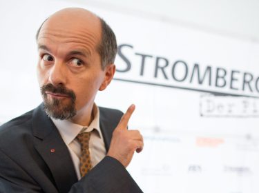 Auf dem Bild zum Stromberg Film 2025 ist Christoph Maria Herbst in seiner Rolle als Bernd Stromberg zu sehen. Er trägt einen dunklen Anzug mit Krawatte und zeigt mit erhobenem Zeigefinger auf den Schriftzug 