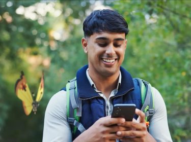 Ein Mann steht im Freien und hat sein Handy in der Hand, während ein Vivilon mit Kontinentalmuster hinter ihm fliegt