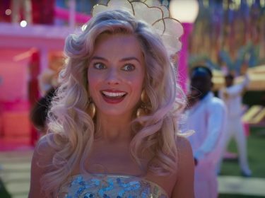 Auf dem Bild zu Barbie 2 ist Barbie, gespielt von Margot Robbie, in einer lebhaften Szene aus dem ersten Barbie-Film zu sehen. Sie trägt ein glitzerndes, mit Pailletten besetztes Kleid und hat voluminöses blondes Haar. Ihr Gesichtsausdruck ist fröhlich und begeistert. Der Hintergrund ist bunt und belebt, mit tanzenden Personen in weißen Outfits und einer festlichen Atmosphäre.