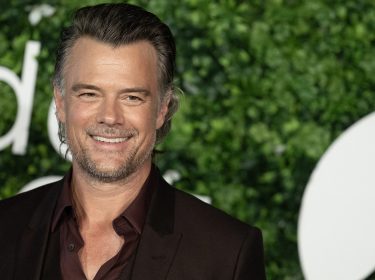 Auf dem Bild ist Josh Duhamel zu sehen, der lächelnd vor einer Kulisse mit grünem Laub und verschwommenem Schriftzug steht. Er trägt einen eleganten dunkelbraunen Anzug mit passendem Hemd und wirkt entspannt und charmant. Das Bild ist Teil einer Präsentation oder Veranstaltung und unterstreicht den Stil des Schauspielers.