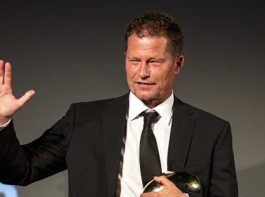 Auf dem Bild zum Film The Neglected ist Til Schweiger zu sehen, der einen schwarzen Anzug mit weißem Hemd und schwarzer Krawatte trägt. Er hebt die rechte Hand lächelnd zum Gruß und hält mit der anderen Hand eine glänzende goldene Trophäe. Der Hintergrund ist dunkel, was den Fokus auf Schweiger legt.