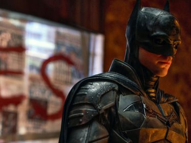 Auf dem Bild zu The Batman 2 ist Robert Pattinson als Bruce Wayne alias Batman zu sehen. Er trägt ein detailliertes, dunkles Batman-Kostüm, das mit Regen bedeckt ist, und blickt ernst zur Seite. Im Hintergrund sind unscharfe Zeitungsseiten mit rotem, aufgemaltem Schriftzug 