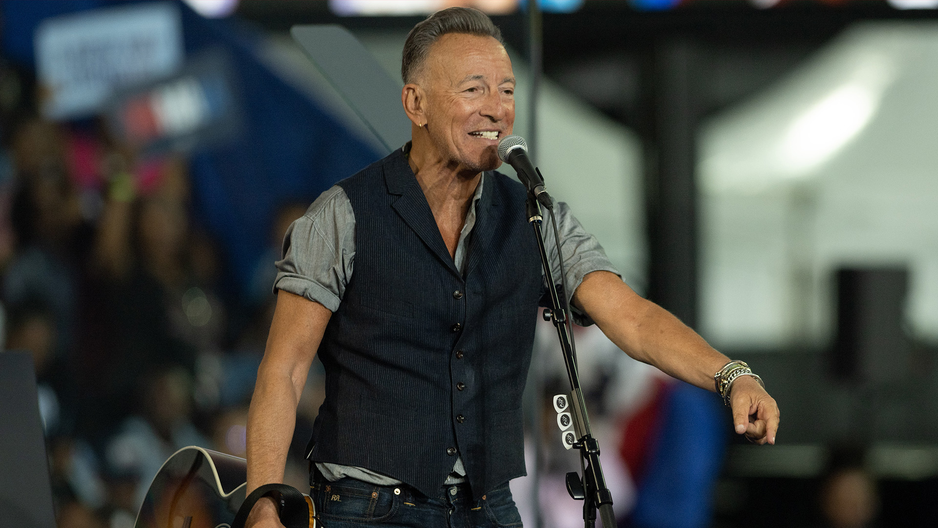 Auf dem Bild in dem Artikel über den Deliver Me From Nowhere-Film ist Bruce Springsteen zu sehen. Er steht auf einer Bühne und trägt ein graues Hemd mit hochgekrempelten Ärmeln sowie eine dunkle Weste. Vor ihm steht ein Mikrofon, und er hält eine Gitarre. Im Hintergrund sind unscharf Zuschauer:innen zu erkennen, die eine lebendige Konzertatmosphäre vermitteln.